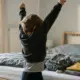 Kind streckt morgens die Arme hoch beim Anziehen, Rucksack und Kleidung liegen bereit