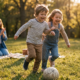 Zwei Kinder rennen lachend einem Ball hinterher, im Hintergrund klatscht ein Elternteil im Park